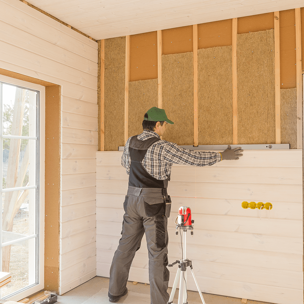 The worker makes finishing works of walls with a white wooden board, using laser line level. Building heat-insulating eco-wooden frame house with wood fiber plates